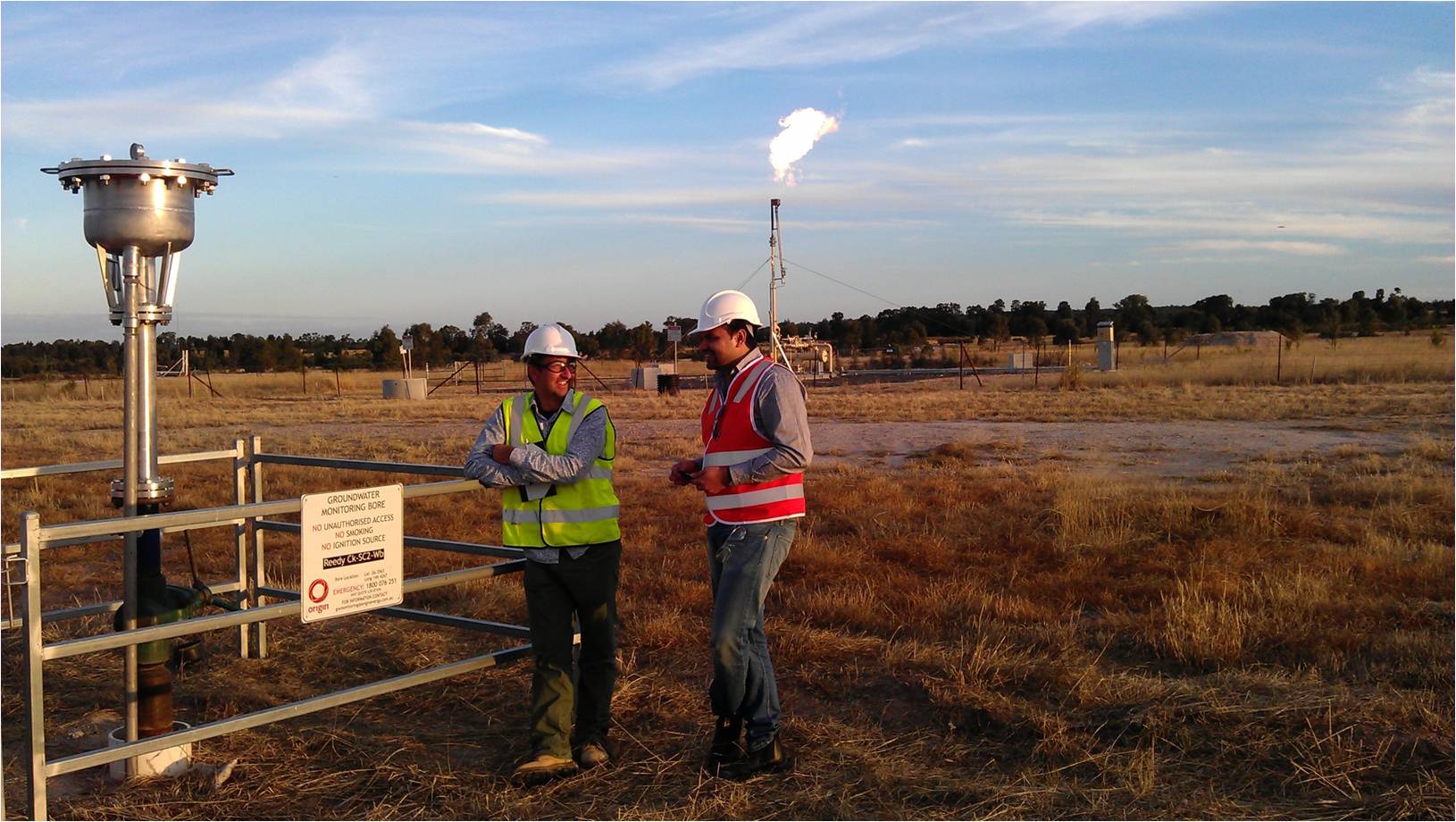 Two men in high visibility vests standing in paddock with bore and gas well